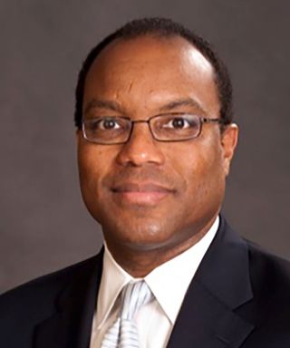 A portrait of Ahmad Corbitt, wearing a black suit and a white-and-blue striped tie, in front of a gray background.