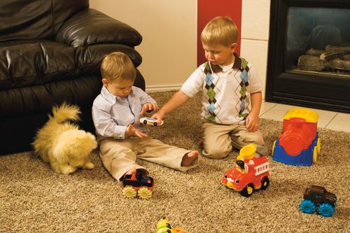 Two little boys play with toy cars while sitting on the living-room floor next to a fireplace and couch.
