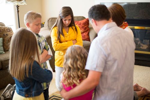 A mother and father kneel down with their four children in their living room and pray together.