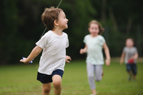 A young child running outside with a kite, with two children running in the background.