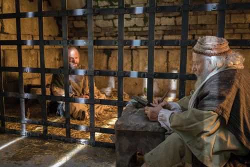 Paul, sitting in a prison cell with his hands shackled, dictating a letter to a scribe.