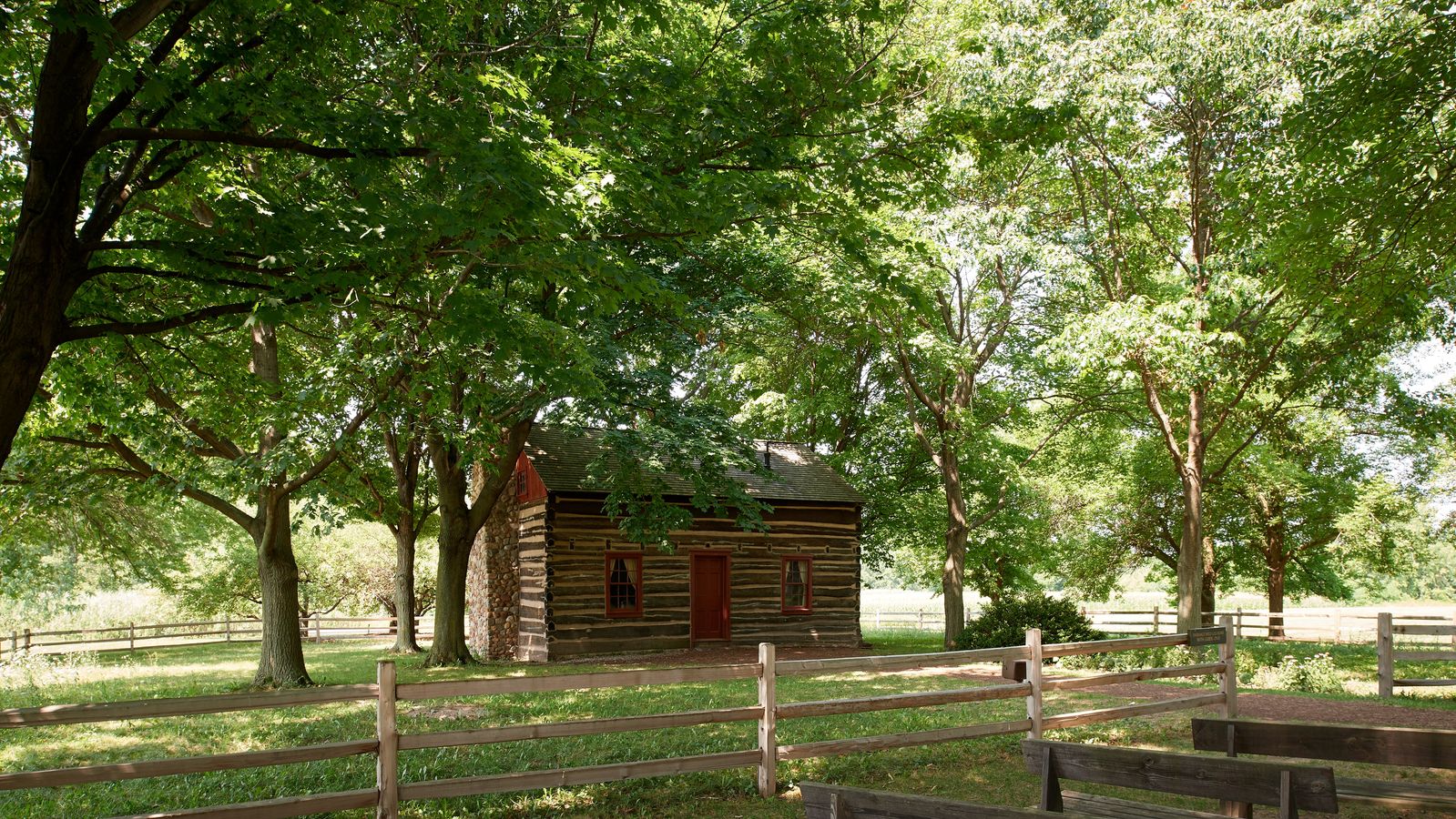 Exterior shots of the Peter and Mary Whitmer Farm. There is a log cabin with a red door. It is surrounded by trees.