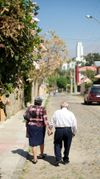 An elderly man and woman holding hands as they walk down a road to the Cochabamba Bolivia Temple in the distance.