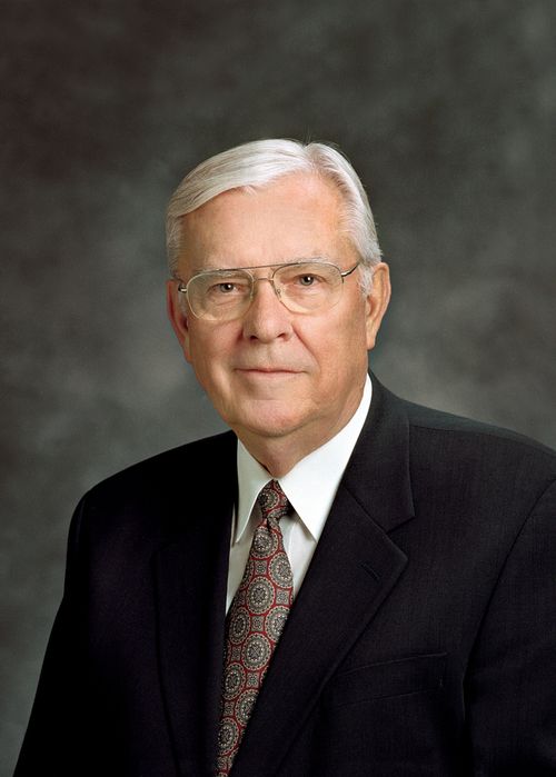A portrait of Elder M. Russell Ballard, who is wearing a black suit and a red and blue patterned tie, in front of a gray background.