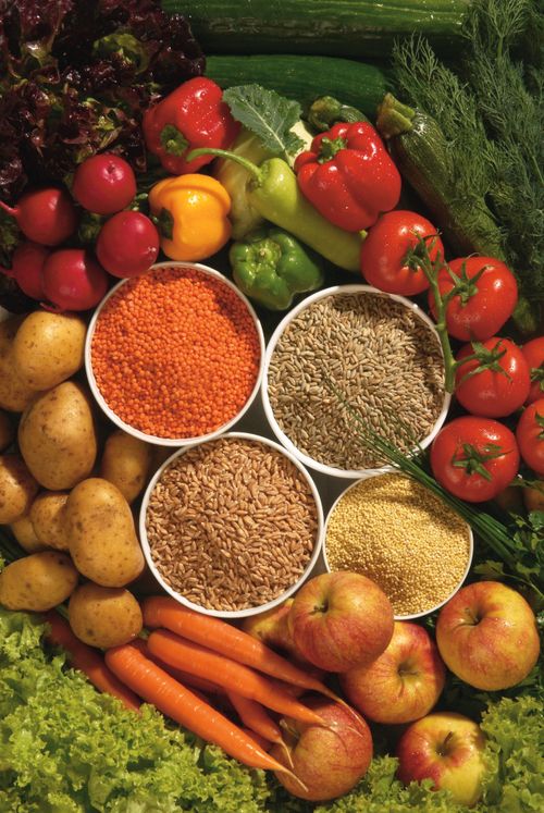 An image of different grains in bowls with peppers, tomatoes, carrots, potatoes, apples, lettuce, and cucumbers around them.