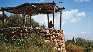 A man standing under the awning of a small stone tower, looking out over the surrounding landscape.