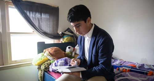 A young man sits on his bed and reads the scriptures. He is dressed in a blue suit and reading in Spanish.