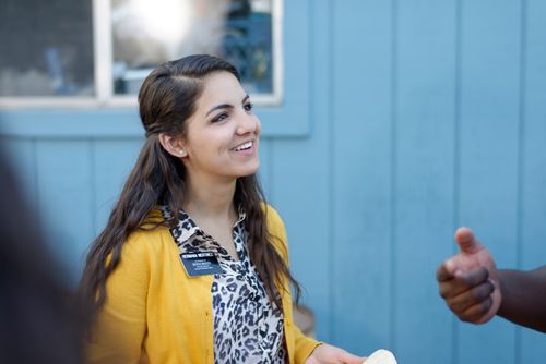 A sister missionary with brown hair and a yellow cardigan smiles as somebody talks to her outside.