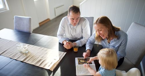 Norway.  Mother and father sitting together in the dining room with their little girl looking at a book, possibly scriptures (Book of Mormon).  Could be having a family home evening.  Daughter pointing at picture of Jesus.