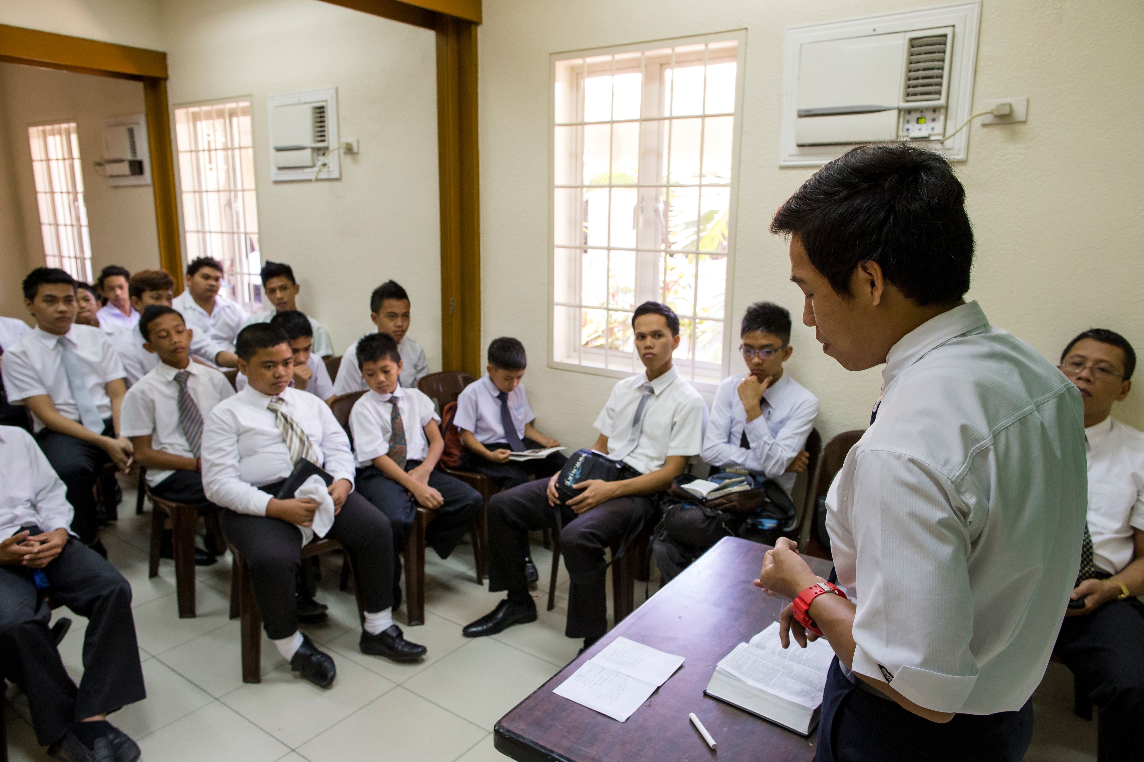 A young man stands to teach a classroom full of young men during a priesthood meeting.  