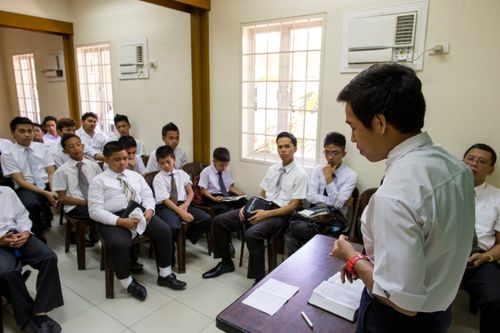 A young man in a white shirt and tie stands to teach a classroom full of young men in white shirts and ties, all sitting down, during a priesthood meeting.