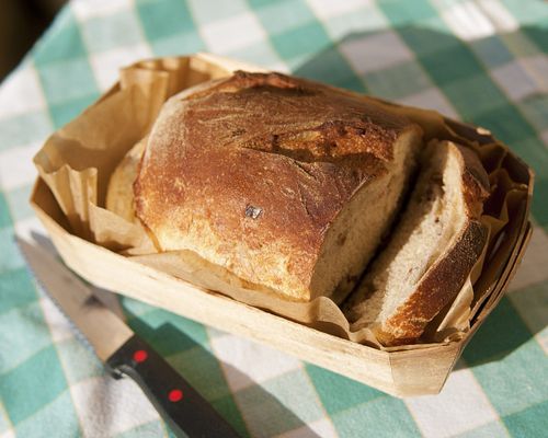 A fresh loaf of sliced bread in a basket lying next to a knife on a green and white plaid tablecloth.