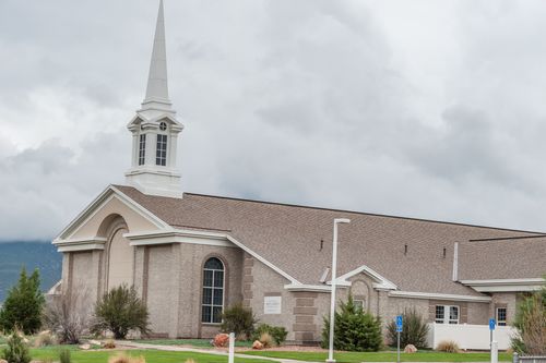 Exterior of light-brown brick, LDS meetinghouse and steeple on a cloudy day.  Official church logo sign can be seen, as well as handicapped parking signs in parking lot. (horiz)