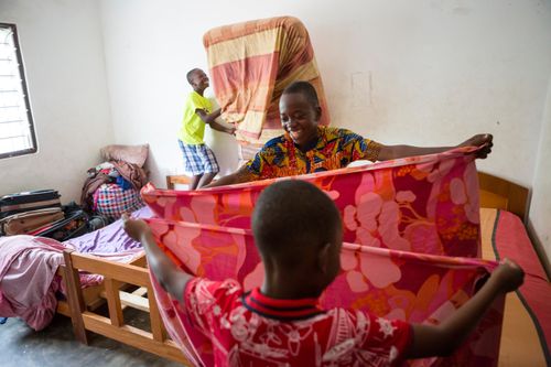 Three brothers in Ghana wearing bright clothing, helping each other with family chores by making beds and folding bedding.