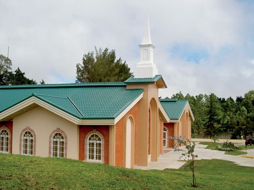 An orange and tan chapel with a green roof, glass windows, and a white steeple, surrounded by a grass lawn in Guatemala.