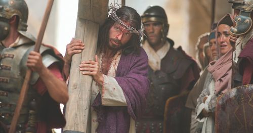 Jesus carries His cross on the way to His Crucifixion while wearing a crown of thorns and purple robe placed on Him by soldiers.