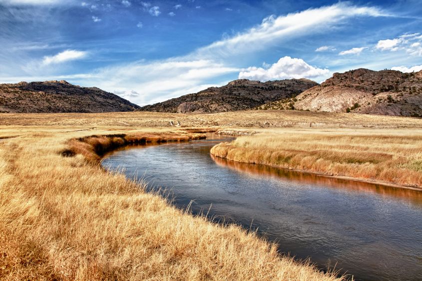 The Sweetwater River runs through a yellow, grassy meadow with hills in the background and clouds overhead.