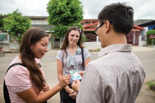 Two dark-haired sister missionaries talking and giving a pass-along card to someone in Thailand.