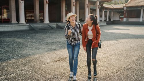 full length two young asian female friends walking visiting in chinese confucius temple talking chatting laughing cheerful. women tourist relax on sunny day with sunset light joyful smiling with joke