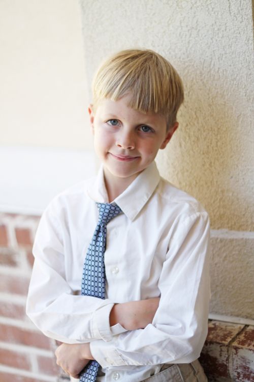 A young boy in a white button-up shirt and tie, folding his arms and smiling.