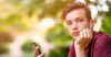 Close-up portrait of a thoughtful unhappy teenage boy with smartphone, outdoors. Sad teenager with mobile phone looks away, in the park. Pensive teenager in casual clothes with cell phone in park