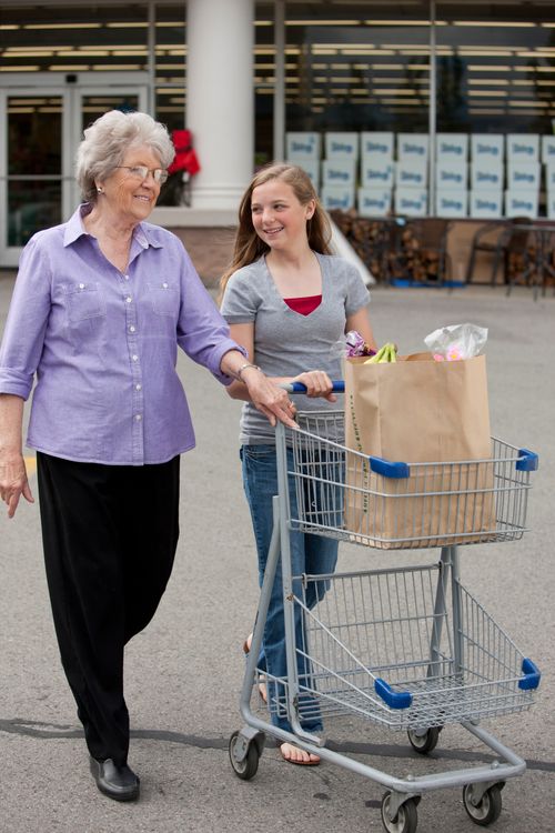 A young woman pushes a shopping cart with a brown bag full of groceries inside while an elderly woman walks beside her.