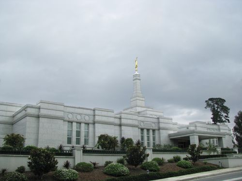The front of the Melbourne Australia Temple, with bushes and small trees growing near the entrance and a dark gray sky overhead.