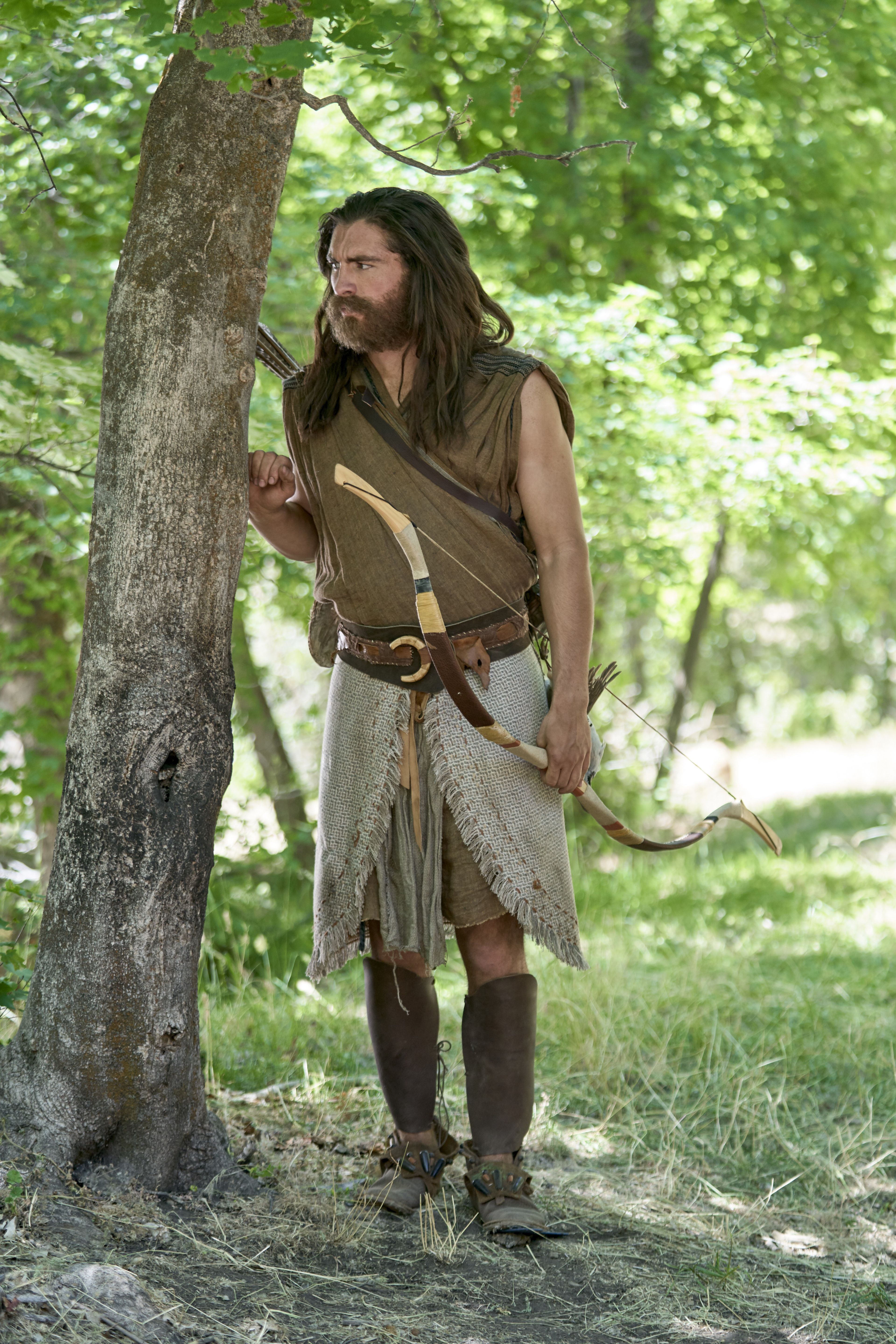 Nephi stands by a tree holding a wooden bow. He overhears his brothers plotting to kill him.
