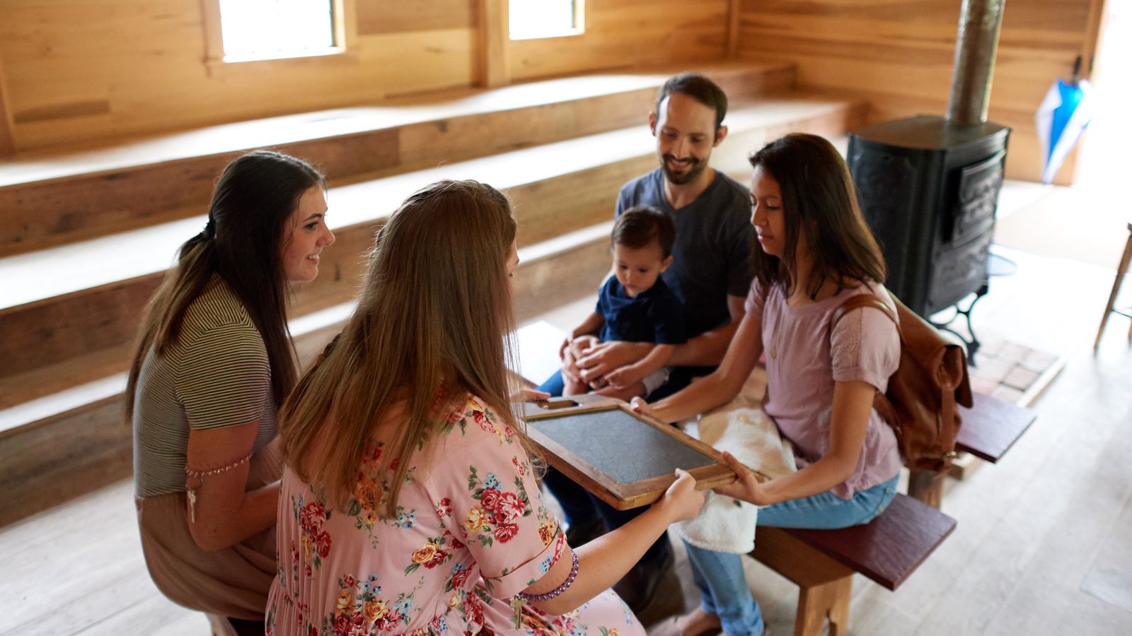A young family sits on a wood bench in a large room with wooden walls. They are instructed by two sister missionaries with chalk slates. 
