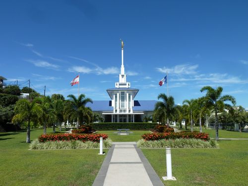 The Papeete Tahiti Temple and grounds on a sunny day, with the Tahitian and French flags flying outside the front of the building.