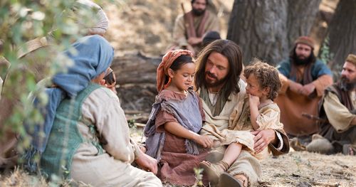 An actor portraying Jesus Christ holding two children on his lap.  There are people sitting near him.
