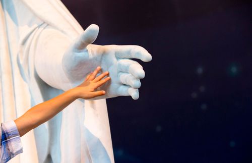 a small child’s hand reaching out and touching the hand of a Christus statue