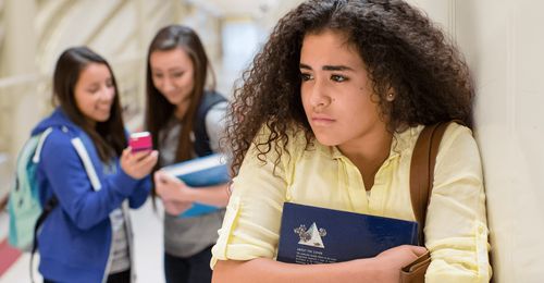 A sad-looking young woman at school.  Behind her are two other young women who are looking at a cell phone and smiling.