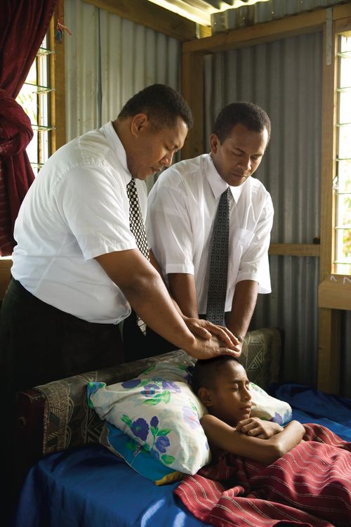 Two men in white shirts and ties give a priesthood blessing to a young boy who is lying sick in bed.