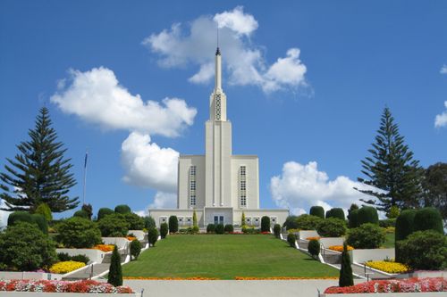 The Hamilton New Zealand Temple on a clear day, with a green lawn and flower beds leading to the entrance.