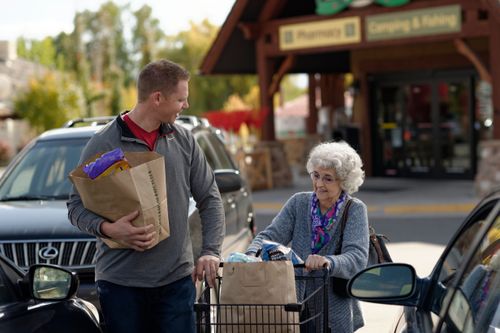 A young man holds a brown bag full of groceries and helps an elderly woman push her cart of groceries out to her car in the parking lot.