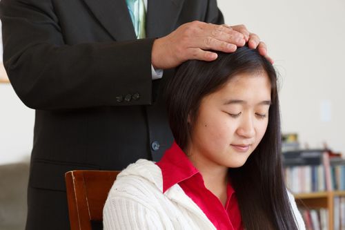 A young woman from Asia receiving a blessing from a Melchizedek Priesthood holder with his hands upon her head.