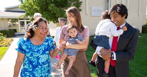 A group of people all walk outside an LDS Meetinghouse. There is a husband, wife, and two sons which they are both holding. Near them is an older Polynesian woman in a blue floral print dress who is smiling and holding the toddlers hand. Behind them is an older couple. Everyone seems to be in conversation and smiling. They are near a white brick LDS Church that has a sign mounted on its front. In front of the building are trees, shrubs, and some flowers.