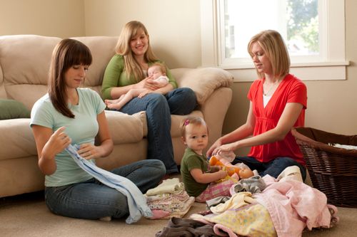 Two women sit on the floor and fold laundry for another woman, who is sitting on the couch with her sleeping baby.