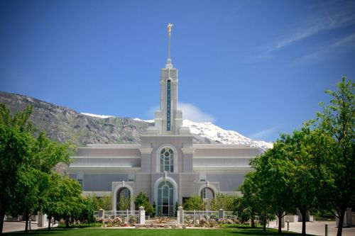 A front view of the Mount Timpanogos Utah Temple, with rows of trees lining the sidewalks and a clear blue sky overhead.
