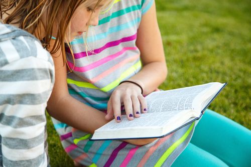 Two young girls in striped shirts sit on the grass and read the Book of Mormon together.