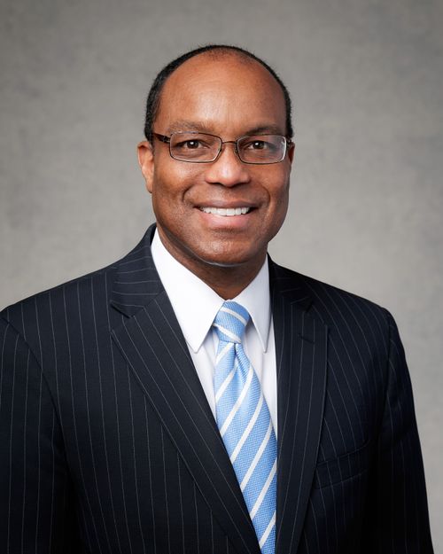 A portrait of Ahmad S. Corbitt wearing a pinstripe suit and a white-and-blue striped tie, in front of a gray background.