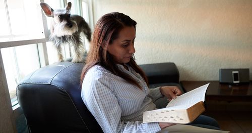Woman reading scriptures on a couch against a window with a dog standing on headrest.