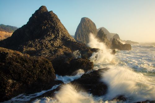 Waves crash onto tall rocks along the Oregon coast with the sun shining down in the distance.