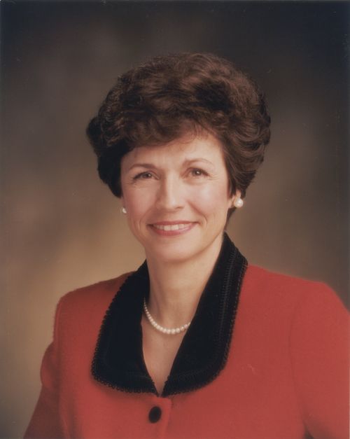 A photograph of Margaret Dyreng Nadauld against a brown background, wearing a red blazer with a black collar.