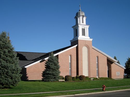 A front view of a brick chapel with a steeple in North Salt Lake, Utah, with grass and pine trees in the front.