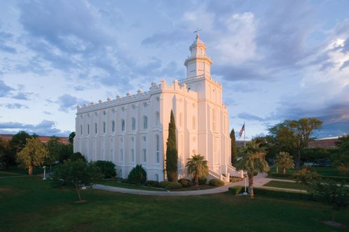 A view of the St. George Utah Temple in the early evening, with the green lawn in the foreground and a U.S. flag near the entrance.