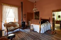 The interior of the Beehive House in Salt Lake City, Utah. This image appears to be a bedroom. There is a bed, a small couch, a wooden chair, and a stove fireplace. There are windows on the sides.