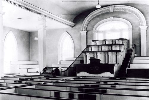 A view inside the Kirtland Temple between 1880 and the 1970s, showing benches, a podium, and large windows.