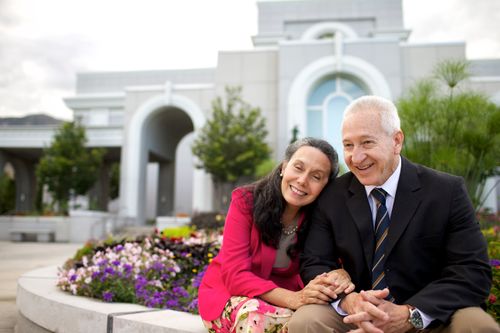 A woman wearing pink and a man in a suit link arms while sitting in front of the Mount Timpanogos Utah Temple.